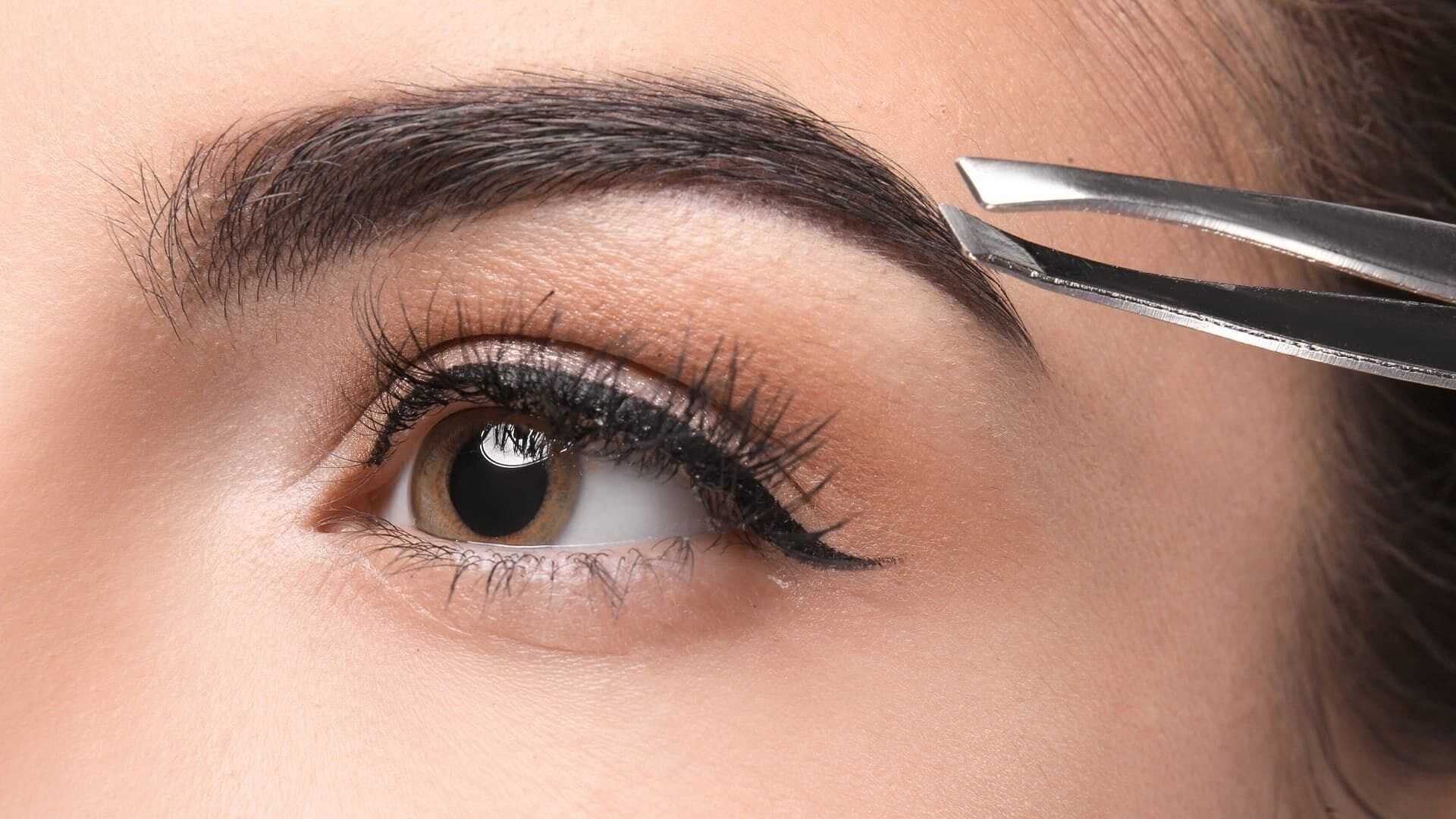 Close-up of an eye with perfectly shaped eyebrows, being groomed with tweezers.