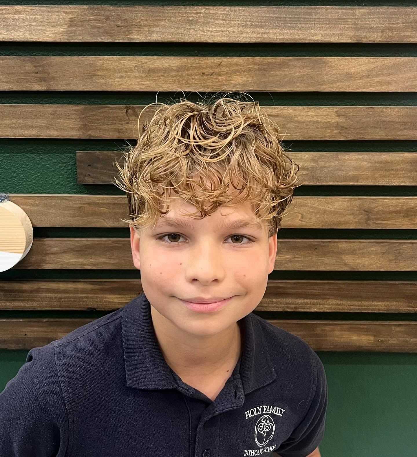 Young boy with curly blond hair smiling, wearing a school uniform in front of a wooden backdrop.