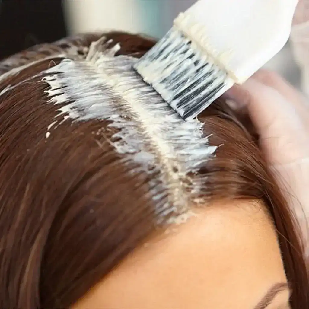 A hairbrush applies dye to brown hair, focusing on the roots for a touch-up.