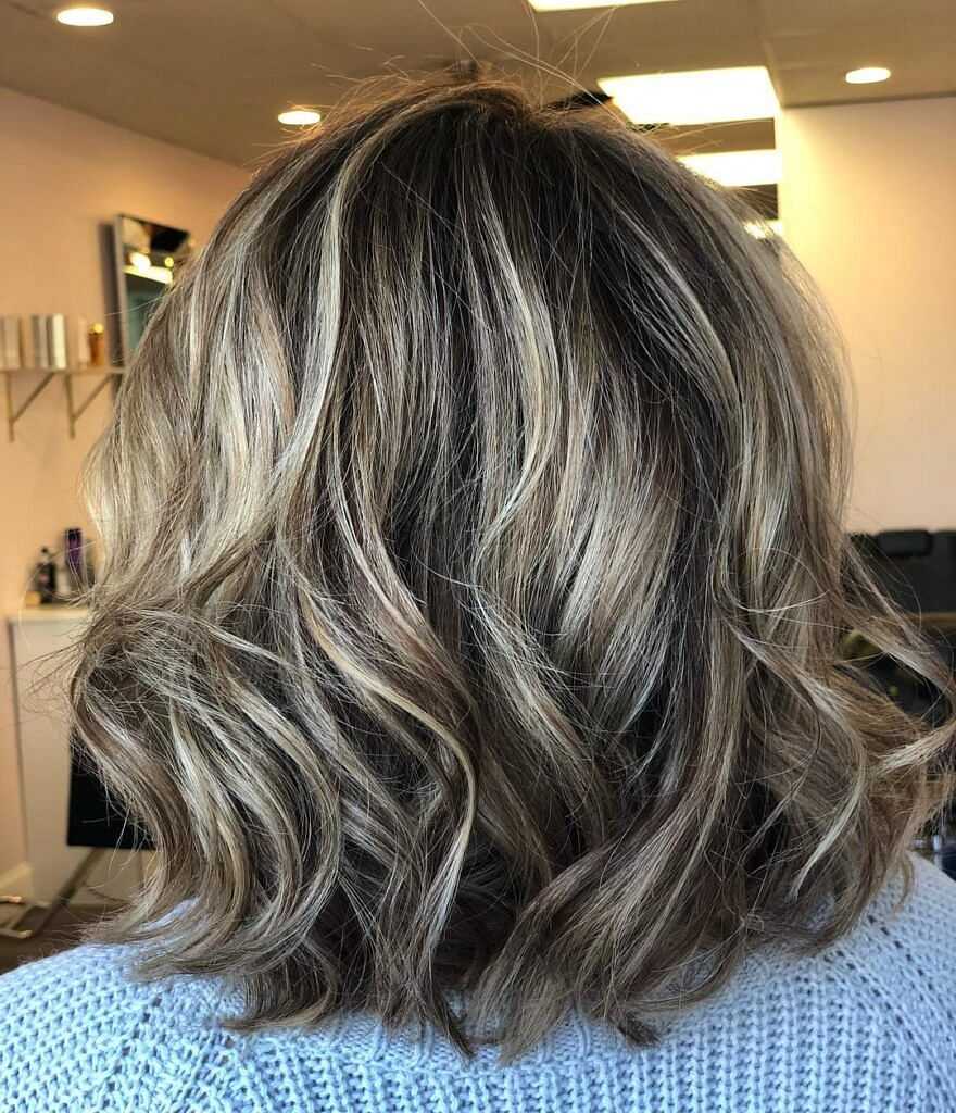 Back view of a woman with shoulder-length, wavy blonde hair and dark roots in a salon.
