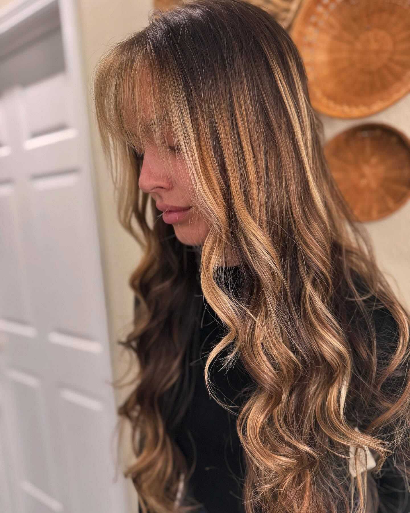 Woman with long, wavy blond hair and bangs, standing in a room with wicker wall decor.