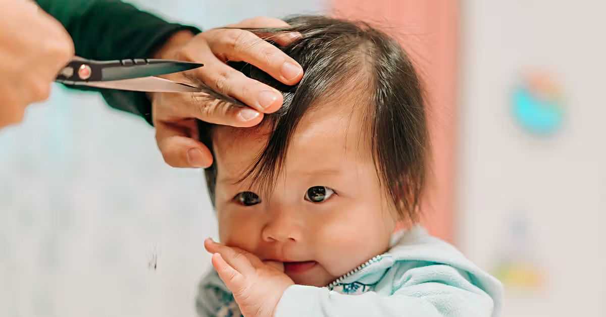 Baby getting a haircut with scissors while sucking thumb, held gently by an adult.